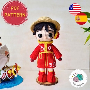 Puede incluir: Patrón de amigurumi de crochet para un personaje rojo y amarillo con un sombrero de paja. El patrón está disponible en inglés y español. El personaje está de pie sobre una base de madera.