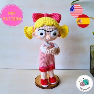 Puede incluir: Muñeca de ganchillo hecha a mano con un vestido rosa, zapatos rojos y un gran lazo rosa. La muñeca tiene el pelo amarillo y está de pie sobre una base de madera. El texto "PDF PATTERN" está en un círculo rosa.