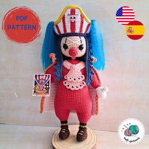 Puede incluir: Muñeca amigurumi de crochet con cabello azul, un sombrero a rayas rojo y blanco y un disfraz de payaso rojo y blanco. La muñeca sostiene un cartel que dice "Circus Bully".