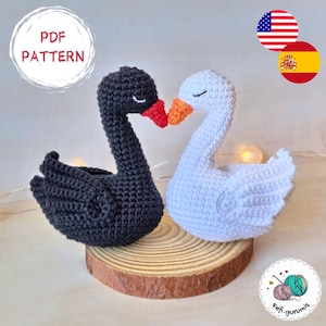 May include: Two crocheted swan figurines, one black and one white, with red and orange beaks, are positioned as if kissing. The swans are on a wooden slice. The text "PDF PATTERN" is in a circle above the swans.