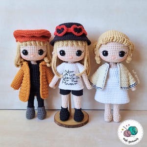 Op de afbeelding: Drie handgemaakte gehaakte poppen met blond haar en grote ogen. De poppen dragen verschillende outfits, waaronder een mosterdkleurige jas, een wit t-shirt met tekst en een witte jurk. Elke pop heeft een unieke hoed.