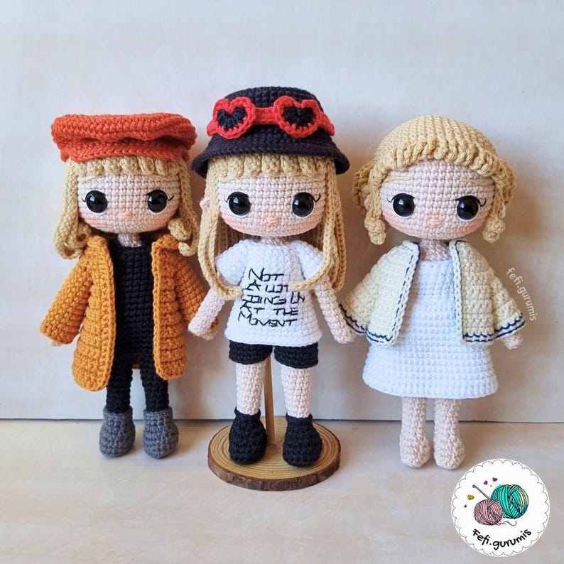 Op de afbeelding: Drie handgemaakte gehaakte poppen met blond haar en grote ogen. De poppen dragen verschillende outfits, waaronder een mosterdkleurige jas, een wit t-shirt met tekst en een witte jurk. Elke pop heeft een unieke hoed.