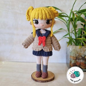 以下が含まれることがあります： 黄色い髪の毛の編みぐるみ人形。茶色のセーター、青いスカート、赤いリボンを着用しています。人形は木の台座の上に立っています。