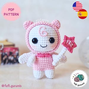 May include: A crocheted doll in shades of pink and white, with a star-shaped wand. The doll has large black eyes and a smiling expression. The text "PDF PATTERN" is in a pink circle. The doll is a charming, handmade design.