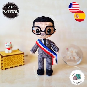 Puede incluir: Una muñeca de ganchillo que representa a una persona con pelo negro, gafas de montura negra y bigote, vestida con un traje gris con corbata negra y una banda en azul, blanco y rojo. La muñeca incluye una pequeña figura de gato blanco.