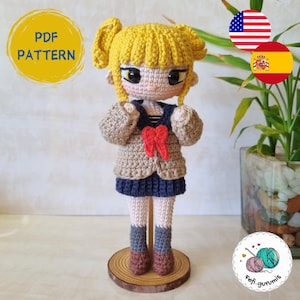 Puede incluir: Muñeca amigurumi de crochet con cabello amarillo, vistiendo un cárdigan marrón, una falda azul y un lazo rojo. La muñeca está de pie sobre una base de madera. La imagen incluye el texto "PDF PATTERN".