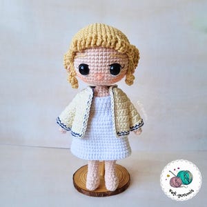 Op de afbeelding: Handgemaakte gehaakte pop met een lichte huidskleur, gekleed in een witte jurk en een cr&egrave;mekleurig jasje met marineblauwe bies. De pop heeft blond haar en grote zwarte ogen. Hij staat op een houten voet.