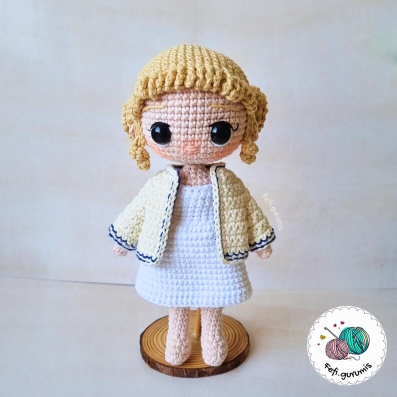 Op de afbeelding: Handgemaakte gehaakte pop met een lichte huidskleur, gekleed in een witte jurk en een cr&egrave;mekleurig jasje met marineblauwe bies. De pop heeft blond haar en grote zwarte ogen. Hij staat op een houten voet.