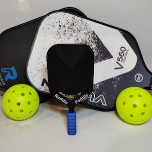 May include: Two black and white pickleball paddles with the text "RONBUS" and "V560 CONTROL" on them. A black pickleball paddle with the text "RADDLE" on it. Three yellow pickleballs.