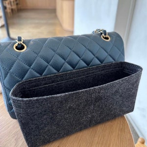May include: A dark gray felt bag insert for a handbag. The insert has a rectangular shape and is made of a soft, textured fabric.