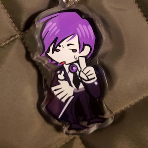 May include: A clear acrylic keyring featuring a cartoon character with purple hair, wearing a black suit and tie. The keyring has a gold-coloured clasp and a gold-coloured ring. The background is a quilted olive green fabric.