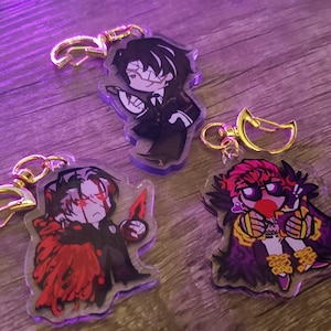 Könnte beinhalten: Drei Acryl-Schlüsselanhänger mit stilisierten Anime-Figuren. Eine Figur trägt einen schwarzen Anzug, eine andere hat rote Akzente und einen dunklen Umhang, und die dritte trägt ein violett-gelbes Outfit mit Brille. Jeder hat einen goldenen Verschluss.