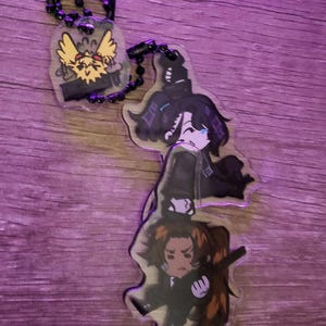 May include: A clear acrylic keyring featuring two stacked chibi-style anime characters with dark hair and dark clothing, connected by a black ball chain to a smaller, separate clear acrylic charm depicting a yellow, spiky-haired creature.