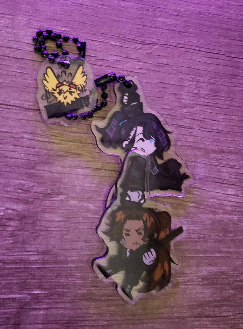 May include: A clear acrylic keyring featuring two stacked chibi-style anime characters with dark hair and dark clothing, connected by a black ball chain to a smaller, separate clear acrylic charm depicting a yellow, spiky-haired creature.