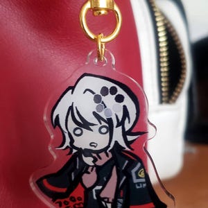 May include: A clear acrylic keychain featuring an anime-style character with white hair and a black and red outfit. The keychain is attached to a gold-colored clasp.