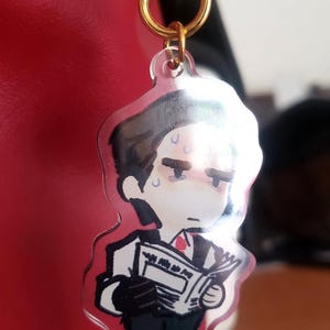 May include: A clear acrylic charm featuring a cartoon illustration of a man in a suit, holding a newspaper. The charm has a gold-colored keychain attachment. The man has a stressed expression.