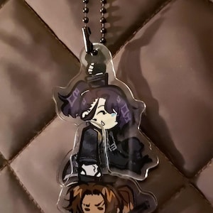 May include: A clear acrylic charm keychain featuring two stacked anime-style characters with dark hair and clothing, suspended from a black ball chain. A small, separate yellow bird-like charm is attached to the top of the chain.