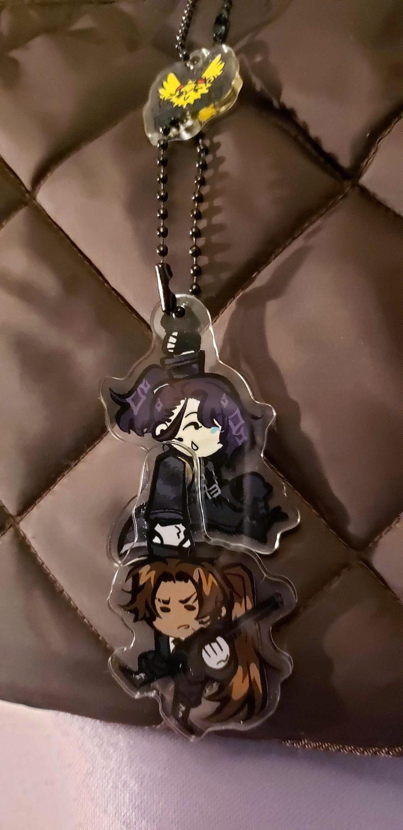 May include: A clear acrylic charm keychain featuring two stacked anime-style characters with dark hair and clothing, suspended from a black ball chain. A small, separate yellow bird-like charm is attached to the top of the chain.