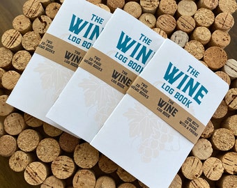 May include: Three white notebooks with the text "The Wine Log Book" printed in blue and brown on the front. The notebooks are stacked on top of each other and have a grapevine design on the front. The notebooks are sitting on a surface covered in cork.