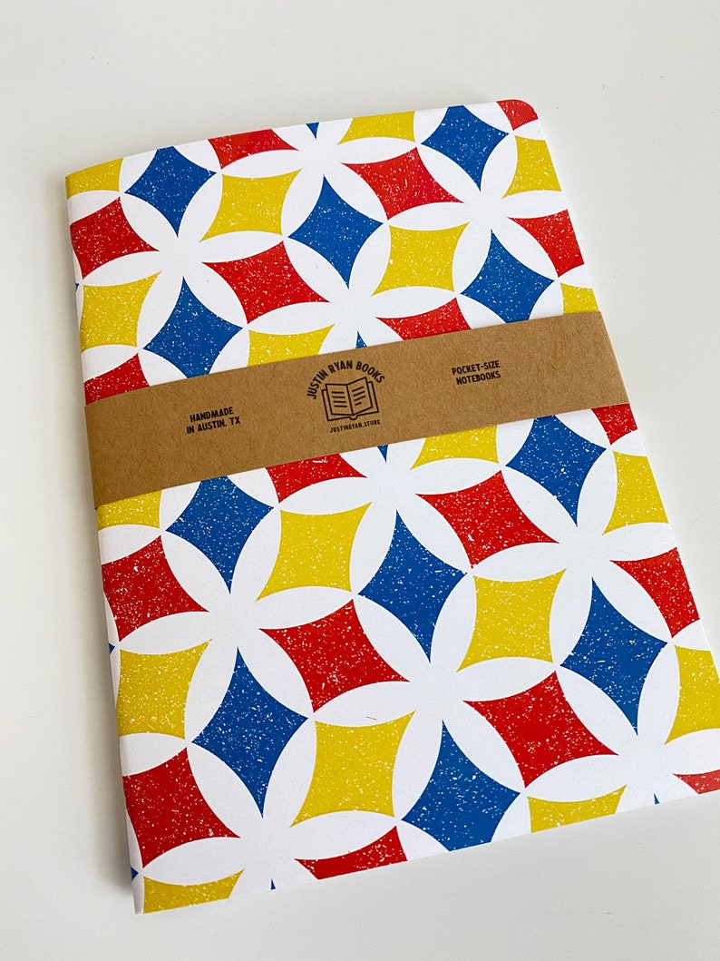 May include: A white notebook with a geometric pattern in red, blue, and yellow. The notebook is handmade in Austin, TX and is pocket-sized. The cover has a brown paper label with the text "Justin Ryan Books" and "Pocket-Size Notebooks".