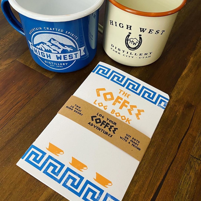 Puede incluir: Un cuaderno blanco con detalles azules y naranjas. La portada presenta un dise&ntilde;o de clave griega y el texto "The Coffee Log Book". El cuaderno est&aacute; envuelto en papel marr&oacute;n con el texto "Log your Coffee Adventures" y "Use both or share with a friend".