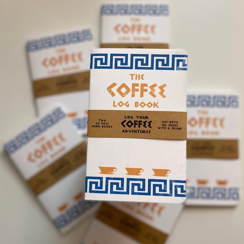 Puede incluir: Dos cuadernos blancos de 20 p&aacute;ginas con dise&ntilde;os de llave griega azul y blanca. La portada del cuaderno presenta el texto "The Coffee Log Book" en naranja y azul. El texto "Log Your Coffee Adventures" est&aacute; impreso en naranja y azul en la portada. Los cuadernos est&aacute;n apilados uno encima del otro.