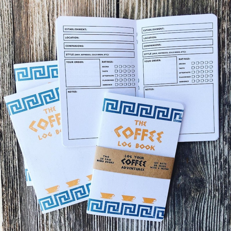 Puede incluir: Dos peque&ntilde;os cuadernos blancos con dise&ntilde;os de llave griega azul y naranja en las portadas. Los cuadernos se titulan "The Coffee Log Book" y presentan un icono de taza de caf&eacute;. El texto "Log Your Coffee Adventures" est&aacute; impreso en una etiqueta marr&oacute;n.