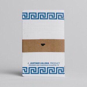 Puede incluir: Un cuaderno blanco con un borde azul de clave griega y una banda de papel marr&oacute;n con un peque&ntilde;o icono de taza de caf&eacute;. El texto "A JUSTINRYAN.ORG PRODUCT DESIGNED AND HANDMADE IN AUSTIN, TX" est&aacute; impreso en el cuaderno.