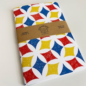 May include: A white notebook with a geometric pattern in red, yellow, and blue. The notebook is handmade in Austin, TX and is a pocket-size notebook. The notebook cover has a brown label with the text "Justin Ryan Books" and "JustinRyan.Store".