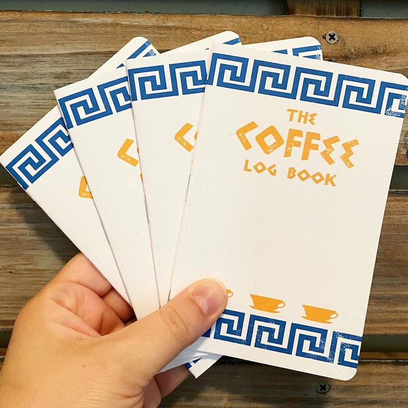 Puede incluir: Cuatro peque&ntilde;os cuadernos blancos con bordes azules y blancos en forma de llave griega. La portada del cuaderno superior dice "The Coffee Log Book" en letras naranjas.
