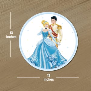May include: A round, light blue graphic featuring Cinderella and Prince Charming. The image shows Cinderella in a blue gown and the Prince in a cream-colored uniform. The graphic is 13 inches in diameter.