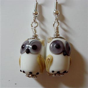 May include: A pair of handcrafted owl earrings. Each earring features a white glass owl with gray and brown accents, black eyes, and silver-colored metal findings. The earrings hang from silver-colored ear wires.