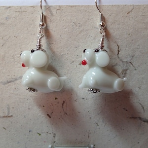 May include: A pair of white glass dog dangle earrings with red accents. The earrings are on a silver hook.