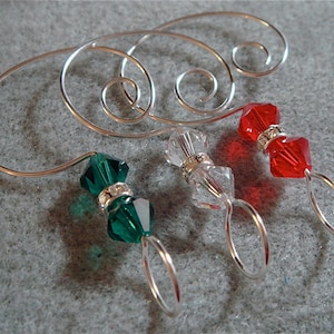 May include: A silver-toned metal knitting stitch marker with a decorative swirl design. Three stitch markers are attached, featuring faceted beads in green, clear, and red, each with a silver ring.