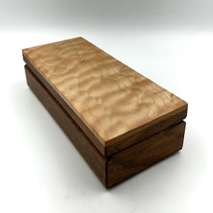 May include: A small wooden box with a light brown top and a dark brown base. The top of the box is made of curly maple wood and the base is made of walnut wood.