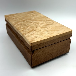 May include: A rectangular wooden box with a light-colored, patterned top and a darker brown base. The box is closed, showcasing the contrast between the two wood tones and the detailed grain of the top.