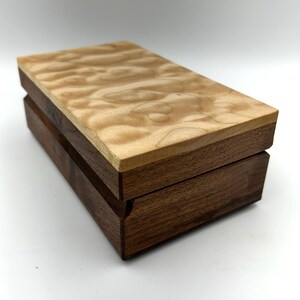 May include: A rectangular wooden box with a light-coloured, patterned top and a dark brown base. The box is made of two different types of wood, with a clear separation between the top and the base. The box is closed and sits on a white surface.
