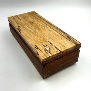 Peut inclure: Une petite boîte en bois avec un couvercle brun clair et une base brun foncé. Le couvercle présente un motif de grain de bois avec des lignes brun foncé.