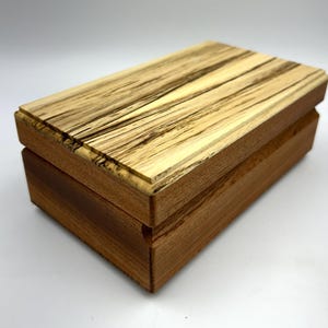 May include: A rectangular wooden box with a lid. The box is made of light brown wood with darker brown streaks on the lid. The box has a simple, classic design, suitable for storing small items.