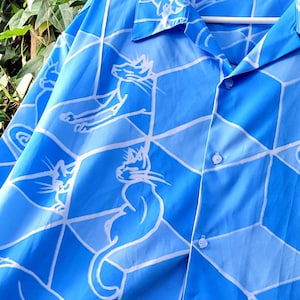 Puede incluir: Camisa azul claro con un dise&ntilde;o geom&eacute;trico blanco e ilustraciones de gatos. La camisa abotonada cuelga de una percha blanca. Fondo de follaje verde.