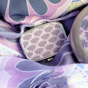 May include: A silver pill box and compact mirror with a fan-shaped design in shades of lavender and gray. The items rest on a lavender and gray patterned scarf. The background includes a patterned fabric in similar colors.