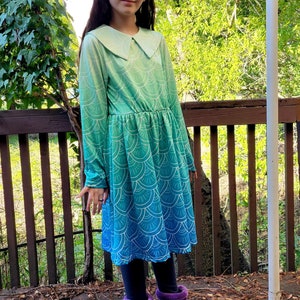 May include: A young person wearing a long-sleeved dress with a green and blue ombre pattern. The dress has a large white collar and a scalloped hem. The person is wearing purple boots.
