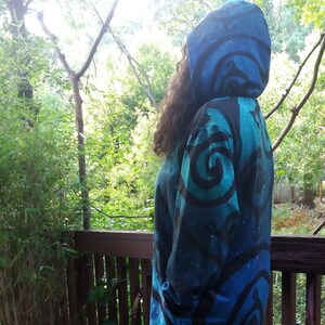 May include: A long-sleeved hooded sweatshirt with a blue and black swirling pattern. The sweatshirt is worn by a person standing on a wooden deck.