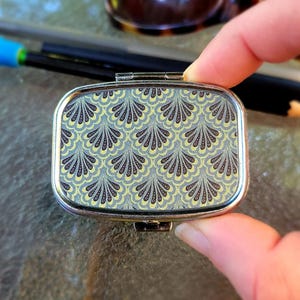 May include: A rectangular, silver-toned pill box with a hinged lid. The top features a repeating pattern of stylized, fan-shaped designs in shades of gray, yellow, and blue. The box is held in a person's hand, with sunglasses and a pen in the background.
