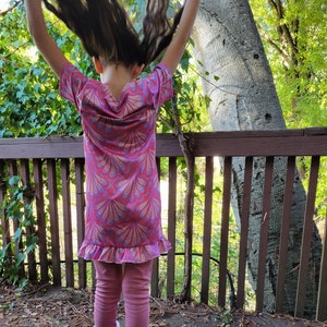 May include: A young person wearing a pink and blue patterned dress with a ruffled hem and pink leggings. They are wearing pink fuzzy boots and have their long brown hair flipped up over their head.