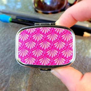 May include: A rectangular, silver-toned pill box with a vibrant pink and light pink fan-shaped pattern. The box is held in a hand, with sunglasses and pencils in the background. The pill box has a hinged lid.
