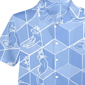 Puede incluir: Camisa azul claro de manga corta con un patr&oacute;n repetido de ilustraciones de gatos blancos sobre un dise&ntilde;o geom&eacute;trico de cubos. La camisa tiene un cuello cl&aacute;sico y cierre de botones.