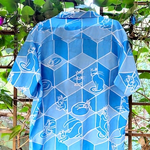 Puede incluir: Camisa abotonada azul claro con un patr&oacute;n geom&eacute;trico de cubos azules e ilustraciones de gatos blancos. La camisa de manga corta tiene cuello y est&aacute; hecha de una tela ligera, perfecta para ropa casual.