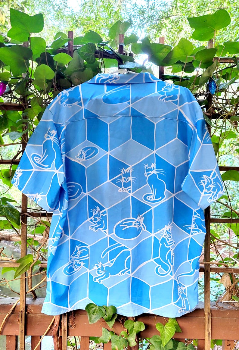 Puede incluir: Camisa abotonada azul claro con un patr&oacute;n geom&eacute;trico de cubos azules e ilustraciones de gatos blancos. La camisa de manga corta tiene cuello y est&aacute; hecha de una tela ligera, perfecta para ropa casual.