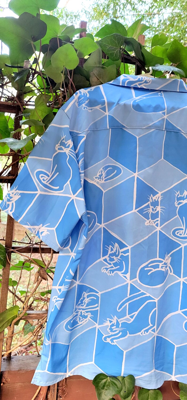 Puede incluir: Camisa azul claro con un patr&oacute;n repetido de ilustraciones de gatos blancos dentro de un dise&ntilde;o geom&eacute;trico. La camisa tiene mangas cortas y est&aacute; hecha de una tela ligera, ideal para el clima c&aacute;lido.